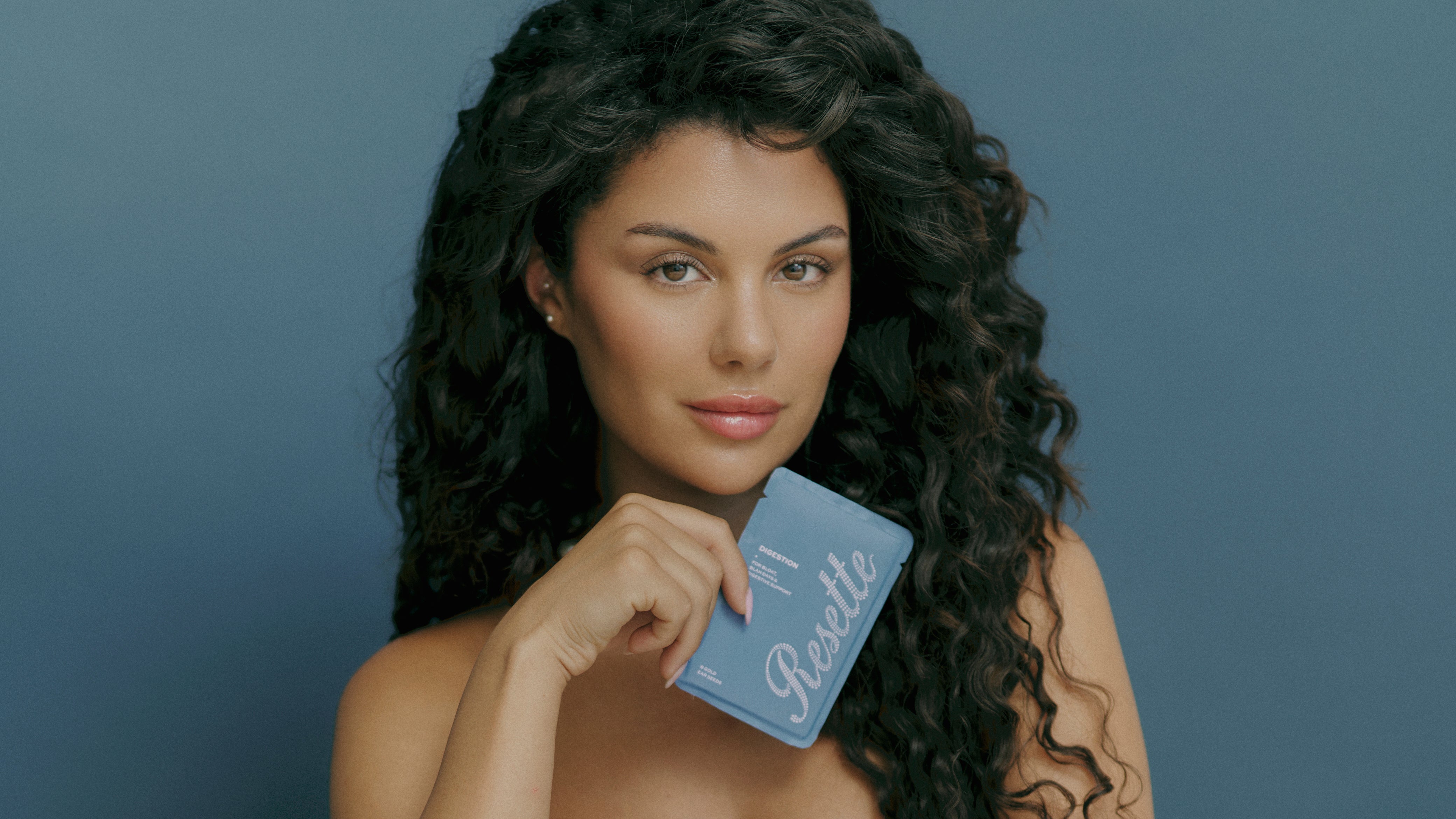 A woman with long, curly dark hair holds a blue digestion Resette Ear Seed sachet against a solid slate blue background.
